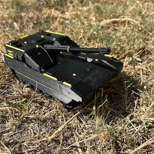 May include: A black toy tank featuring a long main gun barrel and bright yellow accent lines along the chassis, turret, and barrel. The vehicle has a flat, angular design with visible tread tracks on the sides and a matte finish throughout.