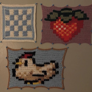 May include: Three rectangular crochet wall hangings. One features a blue and white checkerboard pattern. Another displays a red strawberry with green leaves on a purple background. The third shows a pixelated chicken on a light blue background.