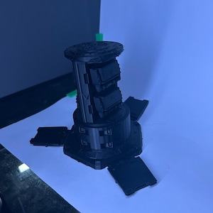 May include: A black 3D-printed object with a cylindrical body and four rectangular panels extending outwards. The top has a detailed, textured design. The object is on a white surface.
