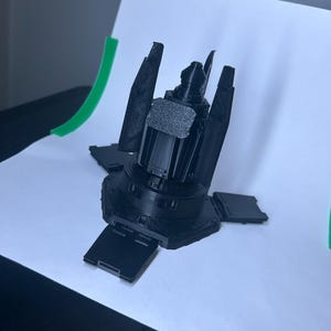 May include: A black mechanical device with a central cylindrical body and several rectangular panels extending outwards. The device has multiple protruding parts and a textured surface. The object is set against a white background.