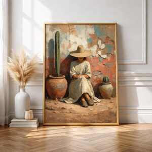 May include: A framed art print depicts a person in a wide-brimmed hat seated near a cactus in a terracotta pot. The background features a textured wall with red, blue, and white paint. The artwork has a warm, earthy color palette.