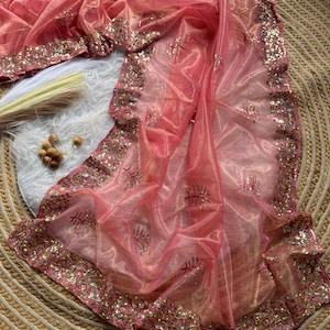 May include: A sheer, pink saree with a gold border and intricate sequin and embroidery detailing. The fabric is draped and folded, showcasing the delicate texture and design. The saree is a traditional garment.