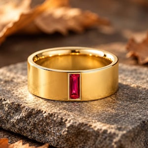 Könnte beinhalten: Ein polierter Goldring mit einem einzelnen, rechteckigen, im Baguetteschliff geschliffenen roten Edelstein, der vertikal in der Mitte gefasst ist. Der Ring hat eine glatte, breite, flache Oberfläche und ein glänzendes Metall-Finish.