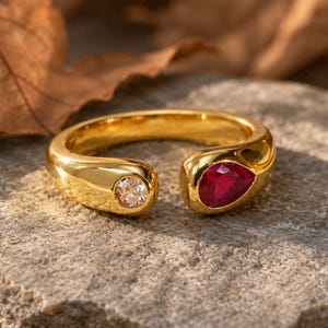 May include: A gold open-style ring featuring a round clear gemstone on one end and a pear-shaped red gemstone on the other. The band has a smooth, polished finish and is designed with an open gap between the two distinct, bezel-set stones.