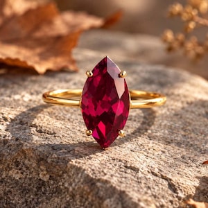 May include: A gold ring featuring a large, marquise-cut, deep red gemstone set in a four-prong mounting. The band is thin and polished, showcasing a vibrant, faceted stone that reflects light across its surface, creating a clear, sharp appearance.