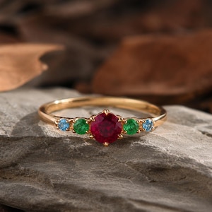 May include: A gold ring with a large, round, red gemstone in the center, flanked by two green and two blue gemstones. The ring is set on a gray stone surface, with blurred brown leaves in the background.