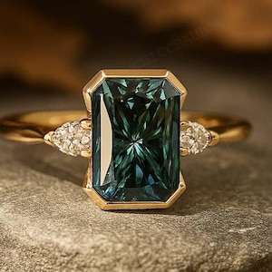 Puede incluir: Un anillo de oro con una gran piedra preciosa de color verde azulado con talla esmeralda. El anillo tiene una banda delicada y pequeñas piedras redondas a cada lado de la piedra principal. El anillo se muestra sobre una superficie de piedra gris.