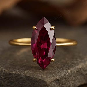 May include: A gold ring with a marquise-cut, deep red gemstone. The ring is set on a dark gray stone surface, with a blurred background. The gemstone is the focal point, showcasing its faceted design and rich color.