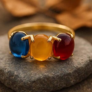 Könnte beinhalten: Ein goldener Ring mit drei ovalen Edelsteinen in Blau, Gelb und Rot. Der Ring liegt auf einem grauen Stein, mit einem verschwommenen Hintergrund in Braun- und Beigetönen. Die Edelsteine sind der Mittelpunkt des Rings.