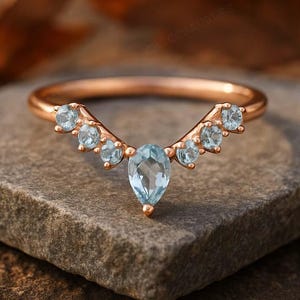 Może przedstawiać: Pierścionek z różowego złota o zakrzywionym kształcie, z niebieskim kamieniem szlachetnym w kształcie łzy pośrodku i mniejszymi okrągłymi niebieskimi kamieniami. Pierścionek leży na kamiennej powierzchni.