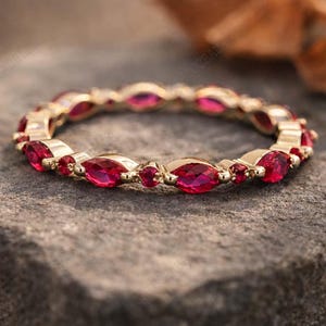 Könnte beinhalten: Ein goldener Eternity-Ring mit abwechselnd im Marquise- und Rundschliff facettierten, leuchtend roten Edelsteinen in einem durchgehenden kreisförmigen Muster. Der Ring ist aus poliertem Gold gefertigt und zeigt ein filigranes Design.