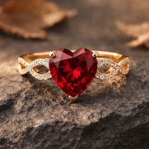 May include: A gold ring featuring a large, faceted heart-shaped red gemstone at the center. The band is designed with an intricate infinity-style twist pattern on both sides, which is encrusted with small, clear, sparkling white accent stones.