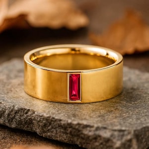 Peut inclure: Une bague en or avec une pierre précieuse rouge rectangulaire au centre. La bague est posée sur une surface en pierre grise, avec des feuilles brunes floues en arrière-plan. La bague a une finition polie et brillante.