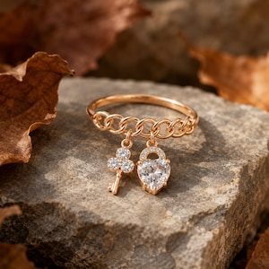 May include: A gold-colored ring with a chain design and two charms: a key and a heart-shaped lock. The key charm has a clover-like design, and the lock charm is encrusted with small, clear stones. The ring is set on a gray stone surface.