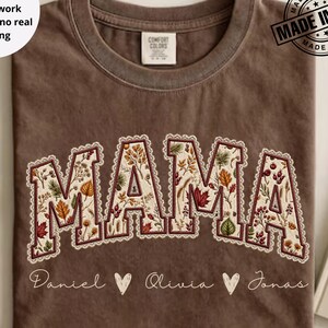 May include: A brown crewneck t-shirt featuring the word MAMA in a large, arched font with a faux patchwork print of autumn leaves and berries. Below the word, the names Daniel, Olivia, and Jonas are printed in white with small heart icons.