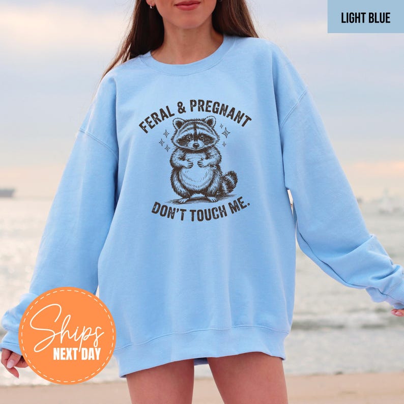A woman wearing a light blue sweatshirt with a graphic of a raccoon and the text "Feral & Pregnant, Don't Touch Me" printed on it.