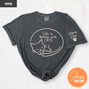 May include: Dark grey t-shirt with a white graphic of a sleeping cat inside an oval, with the text "Life is Better with CATS". A small red heart is above the word "CATS". The sleeve has the text "Charmy Elize" and a paw print.