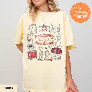 Puede incluir: Una camiseta de color amarillo p&aacute;lido con un estampado gr&aacute;fico que incluye el texto "ER emergency department" en rojo. El dise&ntilde;o presenta varias ilustraciones m&eacute;dicas como un portapapeles, tijeras, un coraz&oacute;n, un monitor y un botiqu&iacute;n.