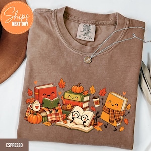 May include: A brown short-sleeve t-shirt featuring a graphic print of anthropomorphic books, pumpkins, and autumn leaves. The books have smiling faces, and one wears glasses and a scarf. A silver necklace rests on the collar of the shirt.