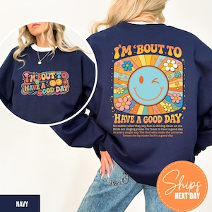 May include: Navy blue sweatshirt with the text "I'M 'BOUT TO HAVE A GOOD DAY" in a retro font. The design includes a smiling sun, flowers, and stars. The back of the sweatshirt has the same design with a longer text.