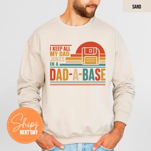 May include: Sand-colored sweatshirt with the text "I KEEP ALL MY DAD JOKES IN A DAD-A-BASE" in a retro design. The graphic includes a floppy disk icon and a sunset-style background. A fun, casual top.