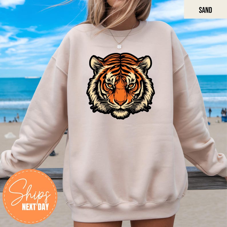 A person wearing a white sweatshirt with a graphic of a tiger's head on it, standing on a beach with a blue sky and ocean in the background.