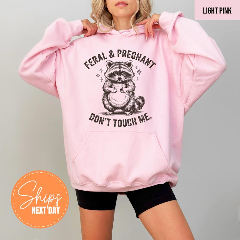 A woman wearing a pink hoodie with a graphic of a raccoon and the text "Feral & Pregnant Don't Touch Me" printed on it.