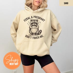 A woman wearing a beige hoodie with a graphic design of a raccoon and the text "Feral & Pregnant, Don't Touch Me."