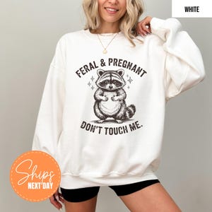 A woman wearing a white sweatshirt with a graphic of a raccoon and the text "Feral & Pregnant Don't Touch Me" printed on it.