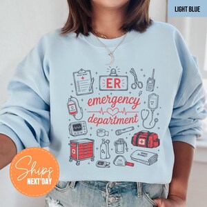 May include: A light blue sweatshirt featuring a graphic print of medical equipment and the text "ER emergency department". The design includes illustrations of a patient report, scissors, a radio, IV bag, stethoscope, medical cart, and a first aid kit.