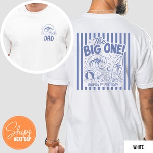 May include: A white t-shirt featuring a blue graphic on the front chest with a wave illustration and the word DAD. The back displays a blue graphic with vertical stripes, a wave, a surfboard, and the text The BIG ONE! RALPH'S 1ST BIRTHDAY.