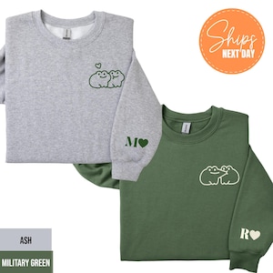 May include: Two sweatshirts, one ash gray and one military green, are displayed. Each has a frog graphic on the chest and a letter with a heart on the sleeve. The ash gray sweatshirt has a green frog graphic, while the military green sweatshirt has a white frog graphic.