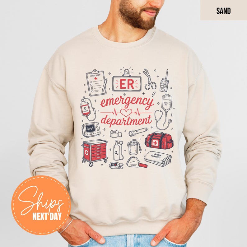 May include: A sand-colored crewneck sweatshirt featuring a graphic print of medical equipment and the text "ER emergency department" with a heart icon. The illustrations include a clipboard, scissors, radio, IV bag, monitor, cart, and oxygen tank.