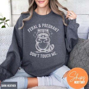 A woman wearing a gray sweatshirt with a graphic of a raccoon and the text "Feral & Pregnant DON'T TOUCH ME."