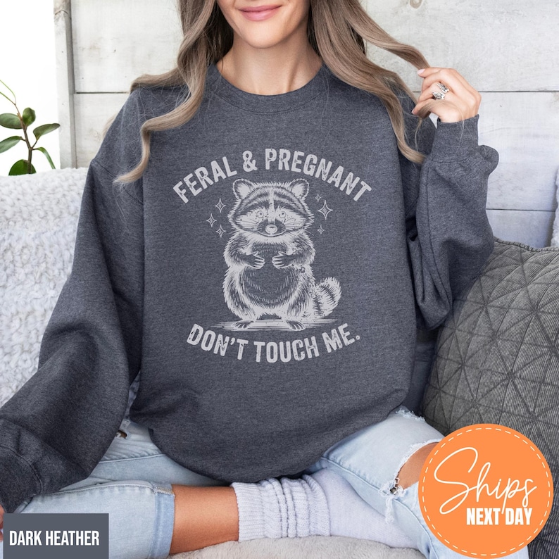 A woman wearing a gray sweatshirt with a graphic of a raccoon and the text "Feral & Pregnant DON'T TOUCH ME."