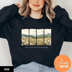 May include: Black sweatshirt featuring a landscape design with white daisies, rolling hills, and mountains. The text "LET KINDNESS BLOOM" is printed below the image. The sweatshirt is a crew neck style.
