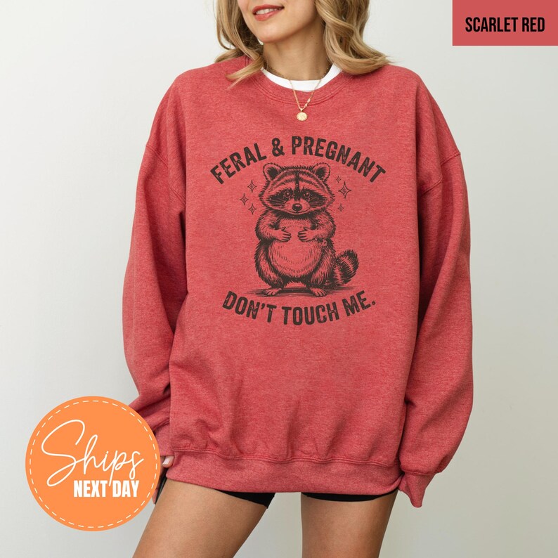 A woman wearing a red sweatshirt with a graphic of a raccoon and the text "Feral & Pregnant, Don't Touch Me" printed on it.