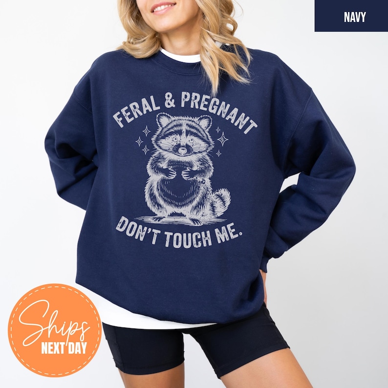 A woman wearing a navy blue sweatshirt with a graphic design of a raccoon and the text "Feral & Pregnant DON'T TOUCH ME."