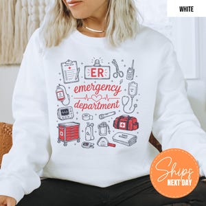 May include: A white long-sleeve sweatshirt featuring a graphic print of medical equipment and the text "ER emergency department". The design includes illustrations of a clipboard, scissors, radio, IV bag, heart monitor, oxygen tank, and medical bag.