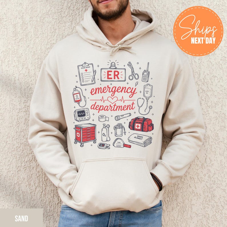 May include: A sand-colored hooded sweatshirt featuring a graphic print of medical equipment and the text "ER emergency department". The design includes illustrations of a clipboard, scissors, heart monitor, oxygen tank, and a red medical bag.