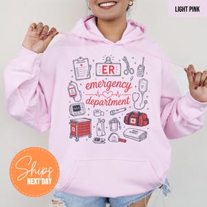 May include: A light pink hooded sweatshirt featuring a graphic print of medical equipment and the text ER emergency department. The design includes illustrations of a clipboard, heart monitor, oxygen tank, medical cart, and first aid kit in red.