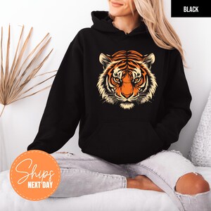 A woman wearing a black hoodie with a tiger graphic on it, sitting on a bed with a palm frond decoration in the background.