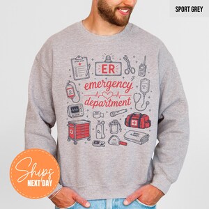 May include: A sport grey crewneck sweatshirt featuring a graphic print of medical equipment and the text "ER emergency department". The design includes illustrations of a clipboard, scissors, a heart monitor, a medical bag, and other supplies.