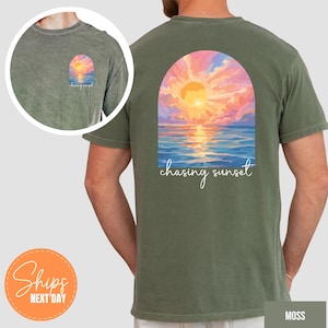 May include: Moss green t-shirt featuring a sunset design. The back of the shirt displays a vibrant sunset over the ocean, framed by an arch, with the words "chasing sunset" in white script. A smaller version of the design is on the front.