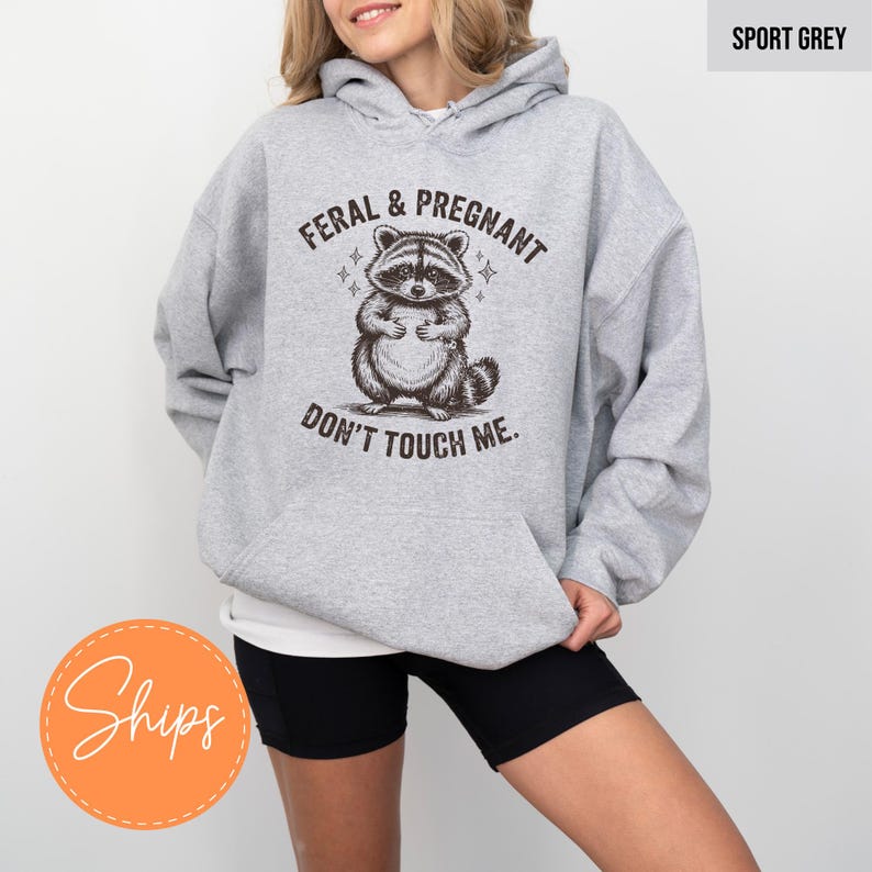A woman wearing a grey hoodie with a graphic of a raccoon and the text "Feral & Pregnant Don't Touch Me" printed on it.