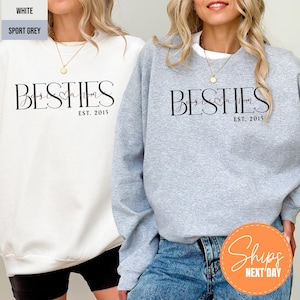 May include: White and sport grey sweatshirts with the word "BESTIES" in black script, with "Boss Mom" and "EST. 2015" below. The sweatshirts are shown on two people. The image also includes a "Ships Next Day" sticker.