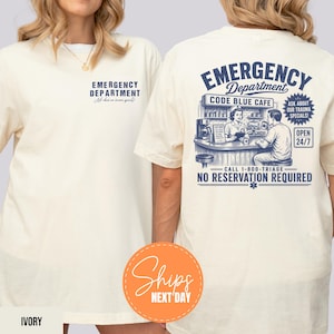 May include: Ivory-colored t-shirt with "EMERGENCY DEPARTMENT" printed on the front. The back features a blue graphic of a cafe scene with text: "CODE BLUE CAFE", "OPEN 24/7", and "NO RESERVATION REQUIRED". A round orange sticker says "Ships NEXT DAY".