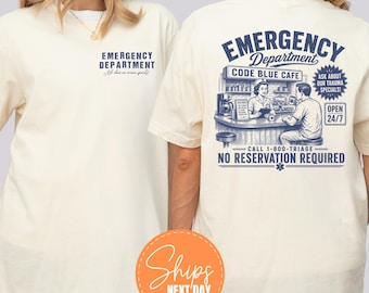 Puede incluir: Una camiseta color marfil con un pequeño gráfico azul en el pecho que dice EMERGENCY DEPARTMENT. La parte posterior muestra un gran gráfico azul de una escena de cafetería llamada CODE BLUE CAFE con el texto EMERGENCY DEPARTMENT y NO RESERVATION REQUIRED.