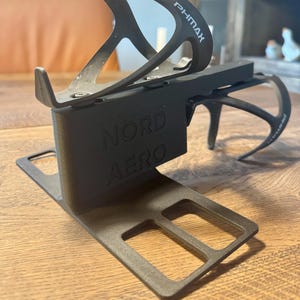 Peut inclure: Support de bidon de vélo noir avec le texte "PHMAX" et "NORD AERO". Le support est conçu pour contenir une bouteille d'eau et est monté sur une base. Le support est fait d'un matériau léger.