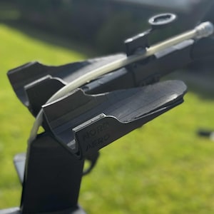 May include: A black aerodynamic bicycle component with the text "NORD AERO" printed on the side. The component has a sleek design with a clear tube running through it. The background is blurred, showing green grass.